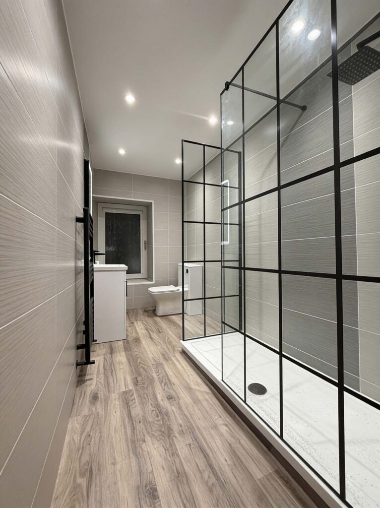 Modern bathroom with walk-in shower featuring black grid glass panels, wood-look flooring, and gray tiled walls